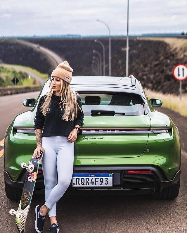 Leticia Bufoni in a black high neck sweater and white pants holding her skateboard in front of her green luxurious car.